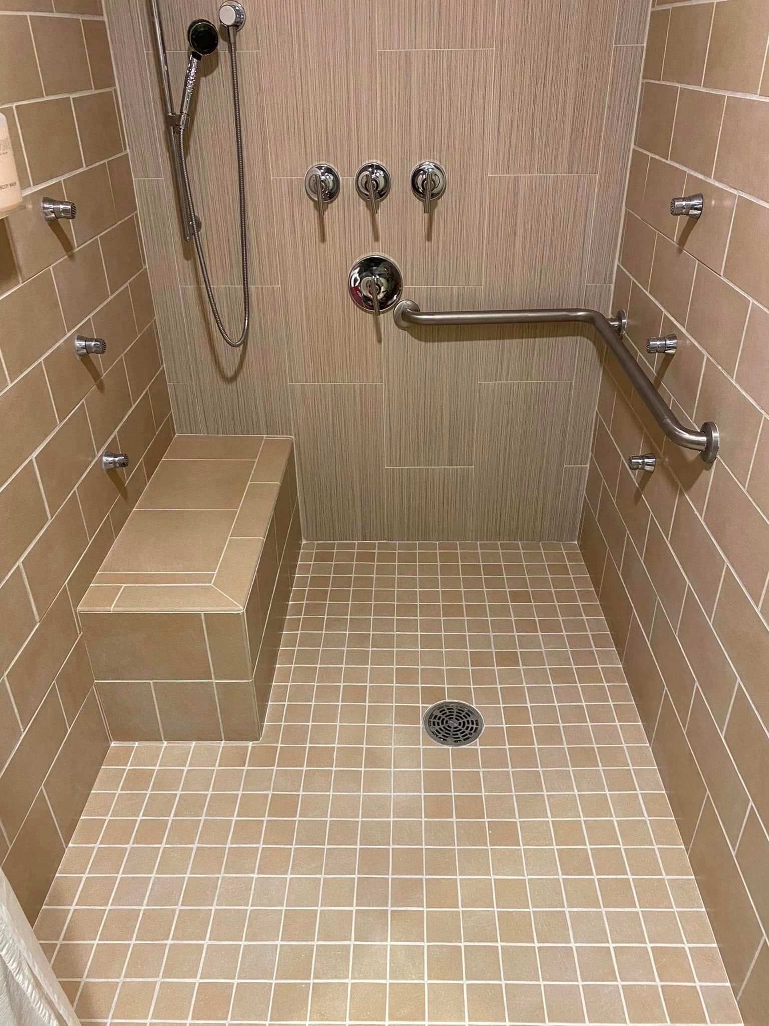 Accessible beige tiled shower with bench, grab bar, multiple shower heads and fixtures.
