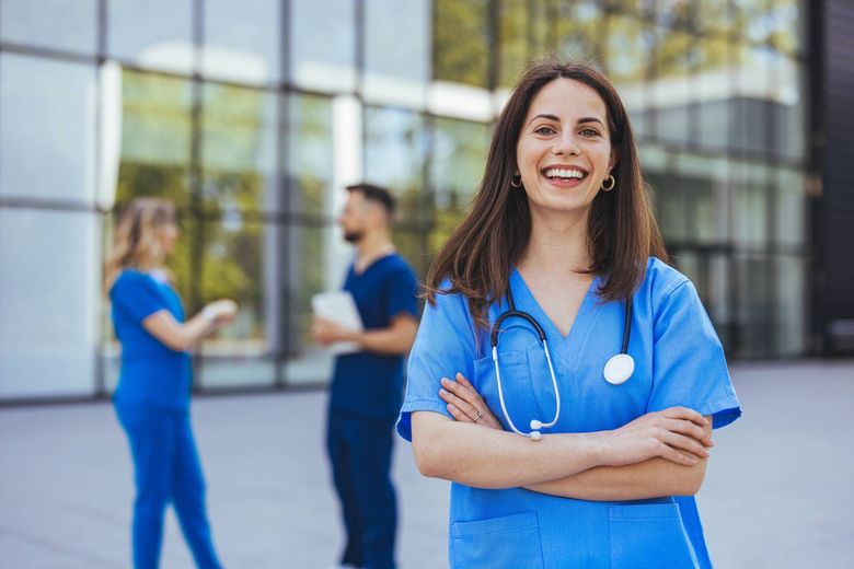 Smiling nurse in blue scrubs with stethoscope standing outside a modern hospital