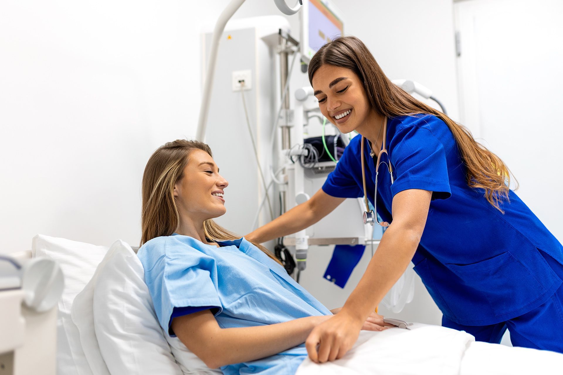 Nurse in blue scrubs smiling and assisting a patient in a hospital bed with medical equipment nearby