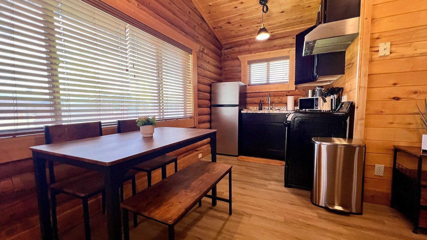 Log cabin interior with kitchen, dining table, and bench. Wooden walls, stainless steel appliances, blinds.