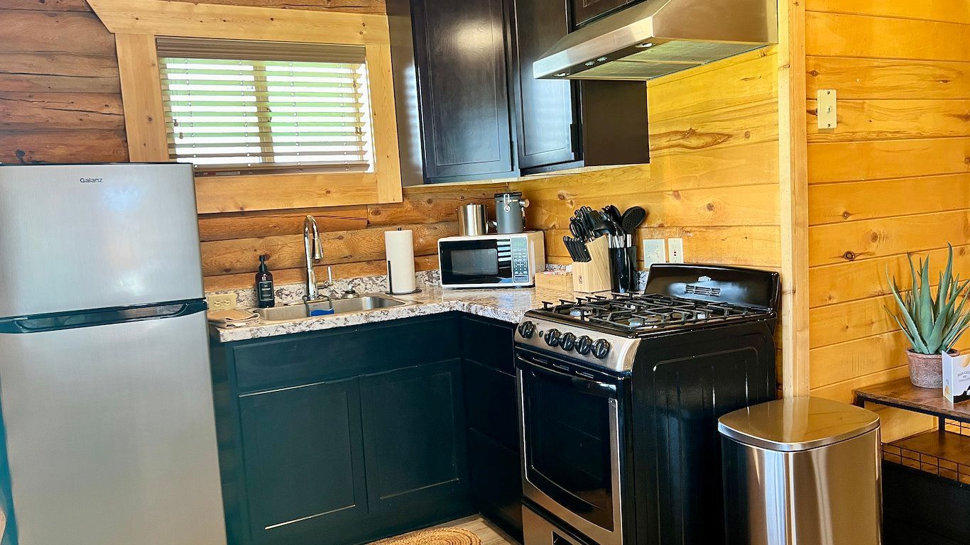 Small kitchen with fridge, cabinets, stove, microwave, and window with blinds. Wooden walls and stainless steel appliances.