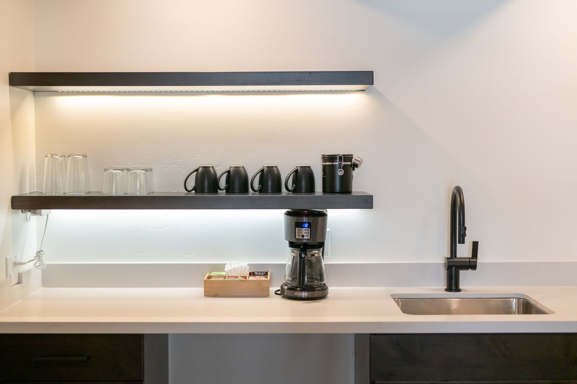 A coffee station with a coffee maker, mugs, glasses, and a sink on a countertop.