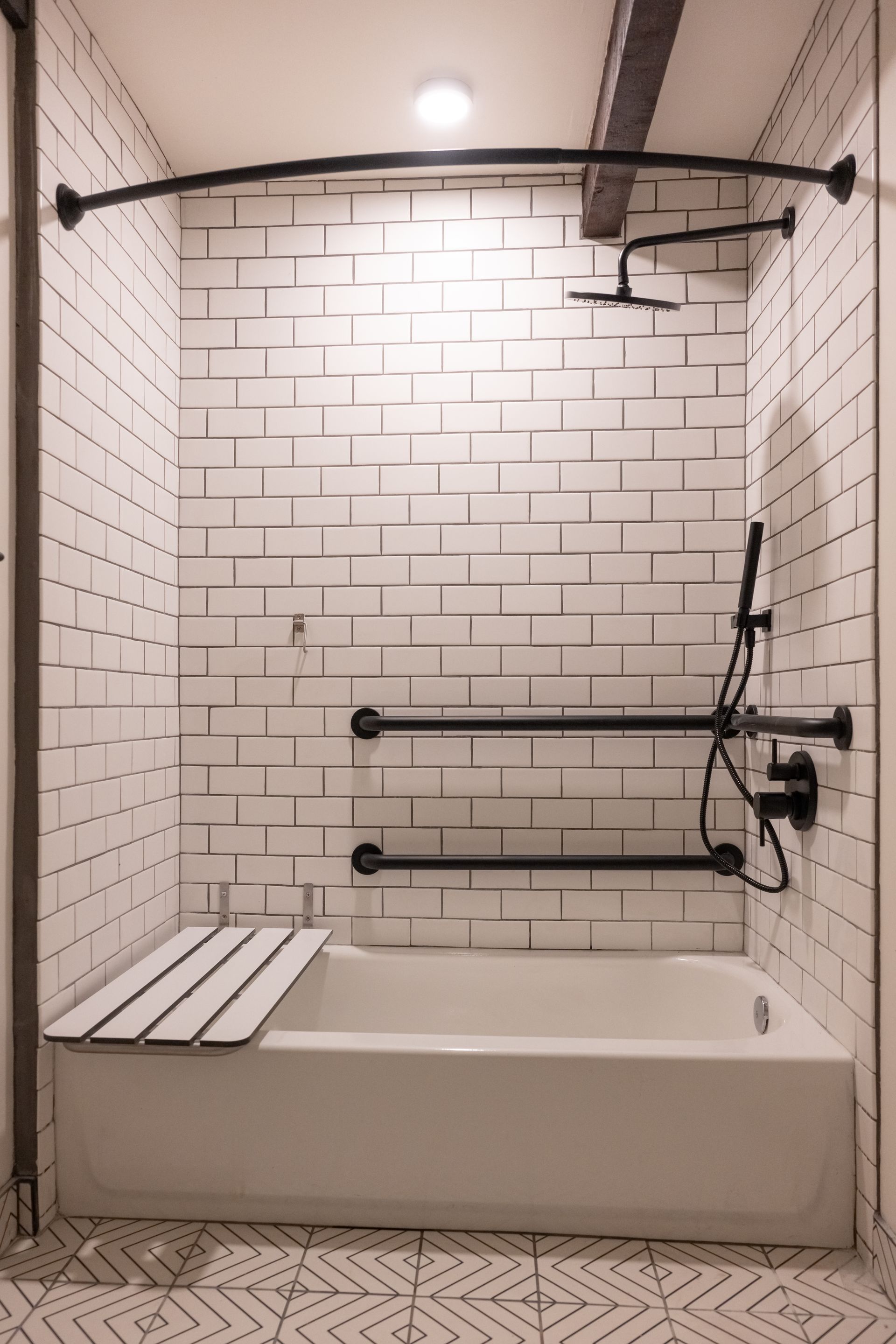 White-tiled shower with black fixtures, curved shower rod, and safety bars over a bathtub with a built-in bench.