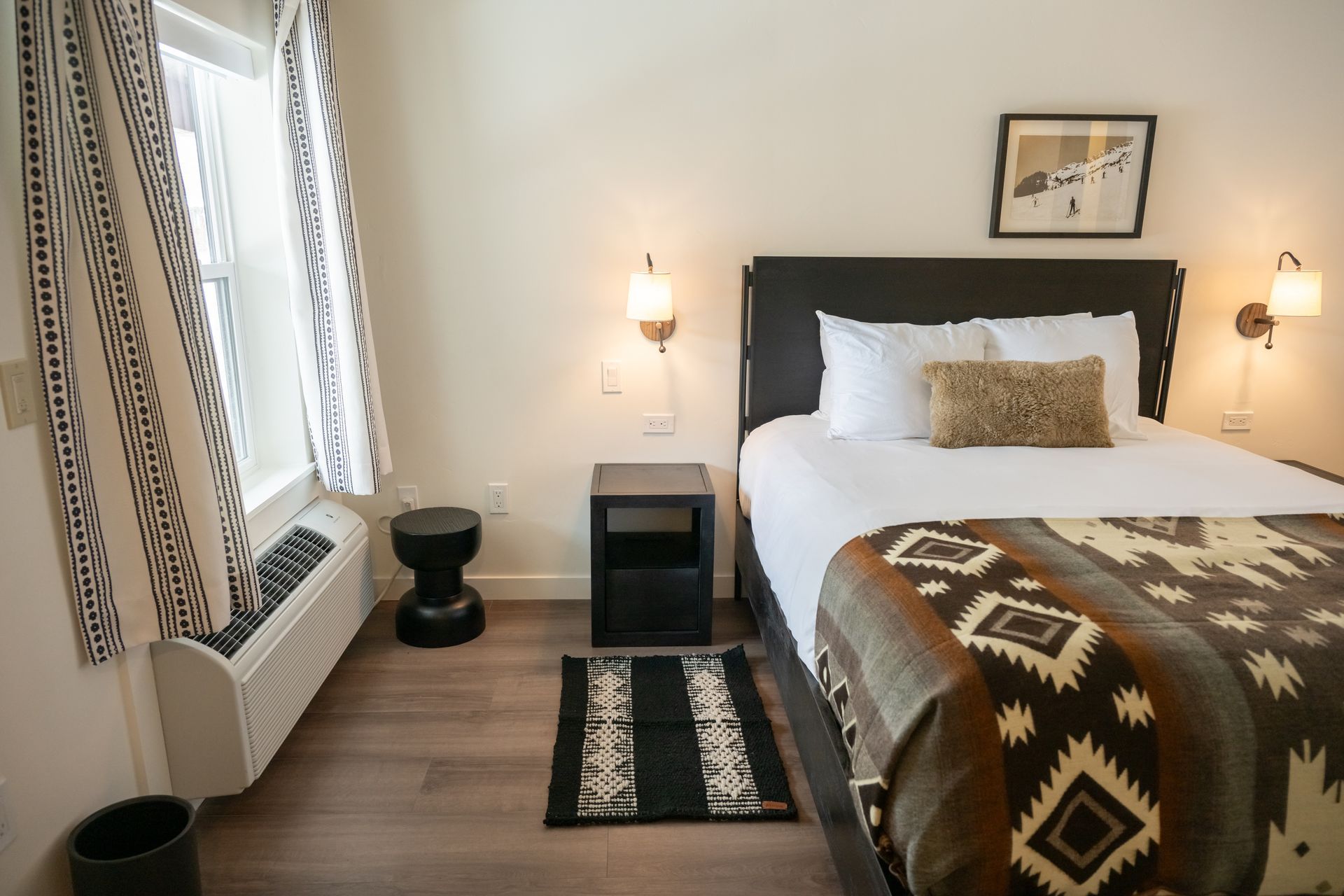 Bedroom with a bed, patterned blanket, window, black nightstand, and wall sconces.