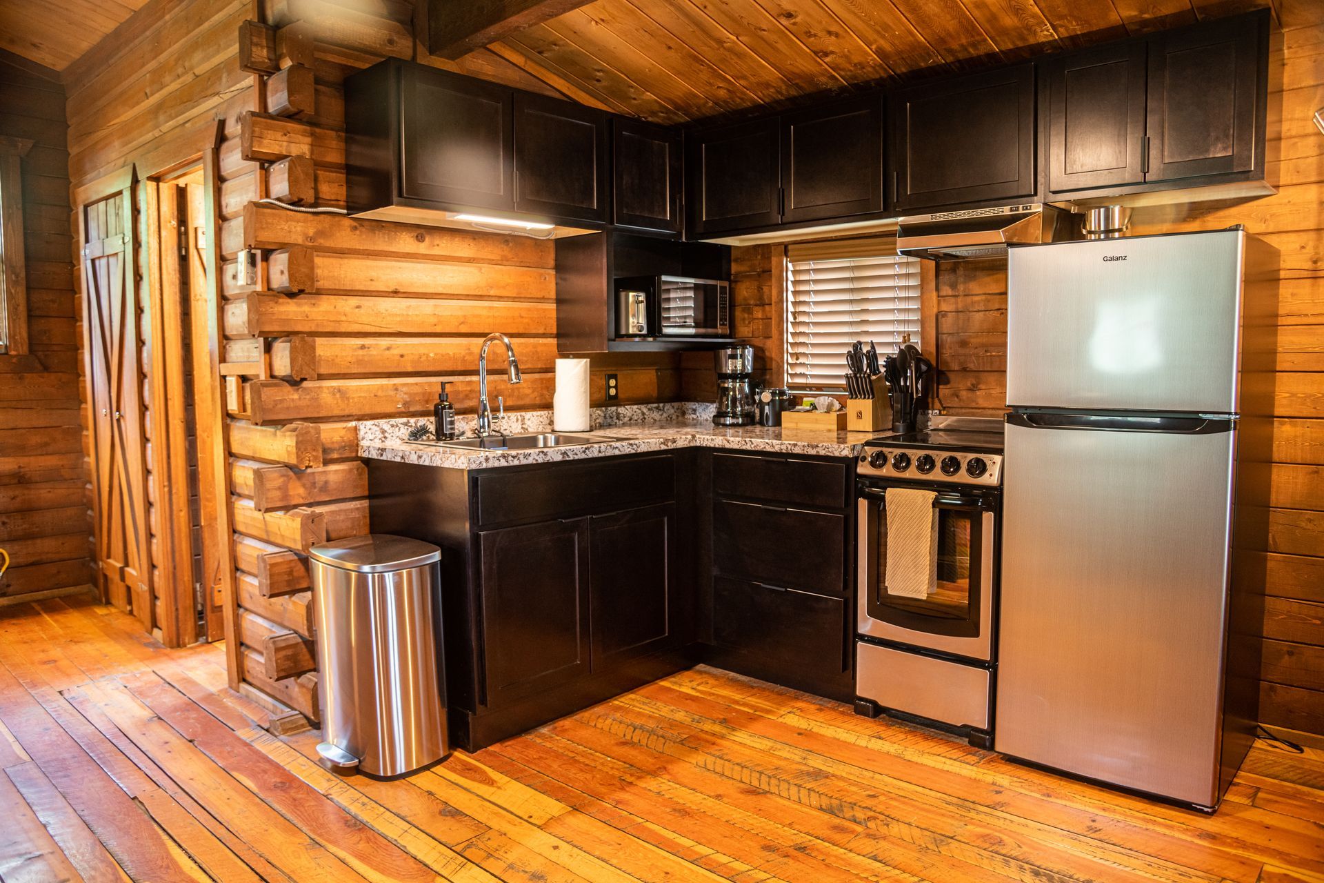 Log cabin kitchen with dark cabinets, stainless steel appliances, and wood walls and floor.