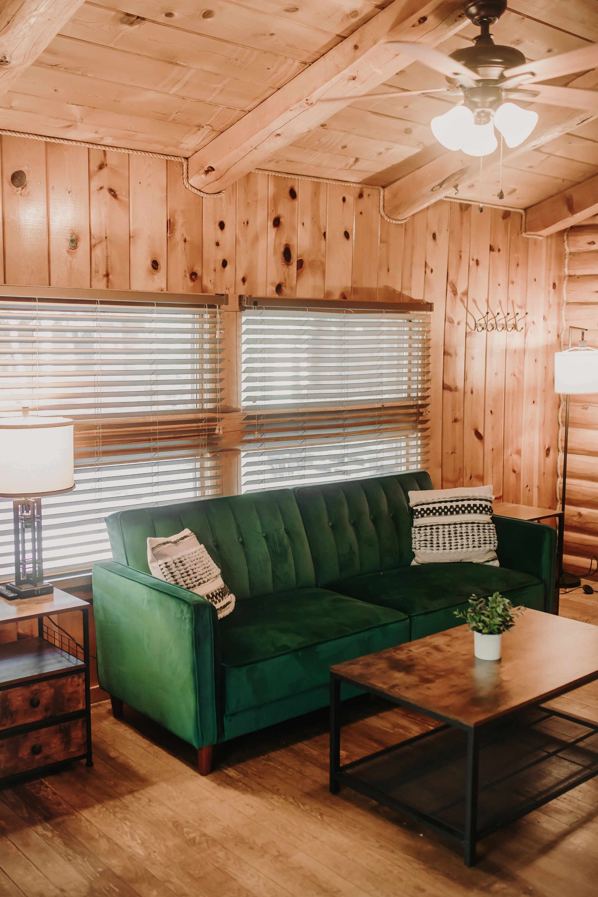 Cozy cabin living room with green velvet sofa, wood paneling, blinds, and small table with plant.