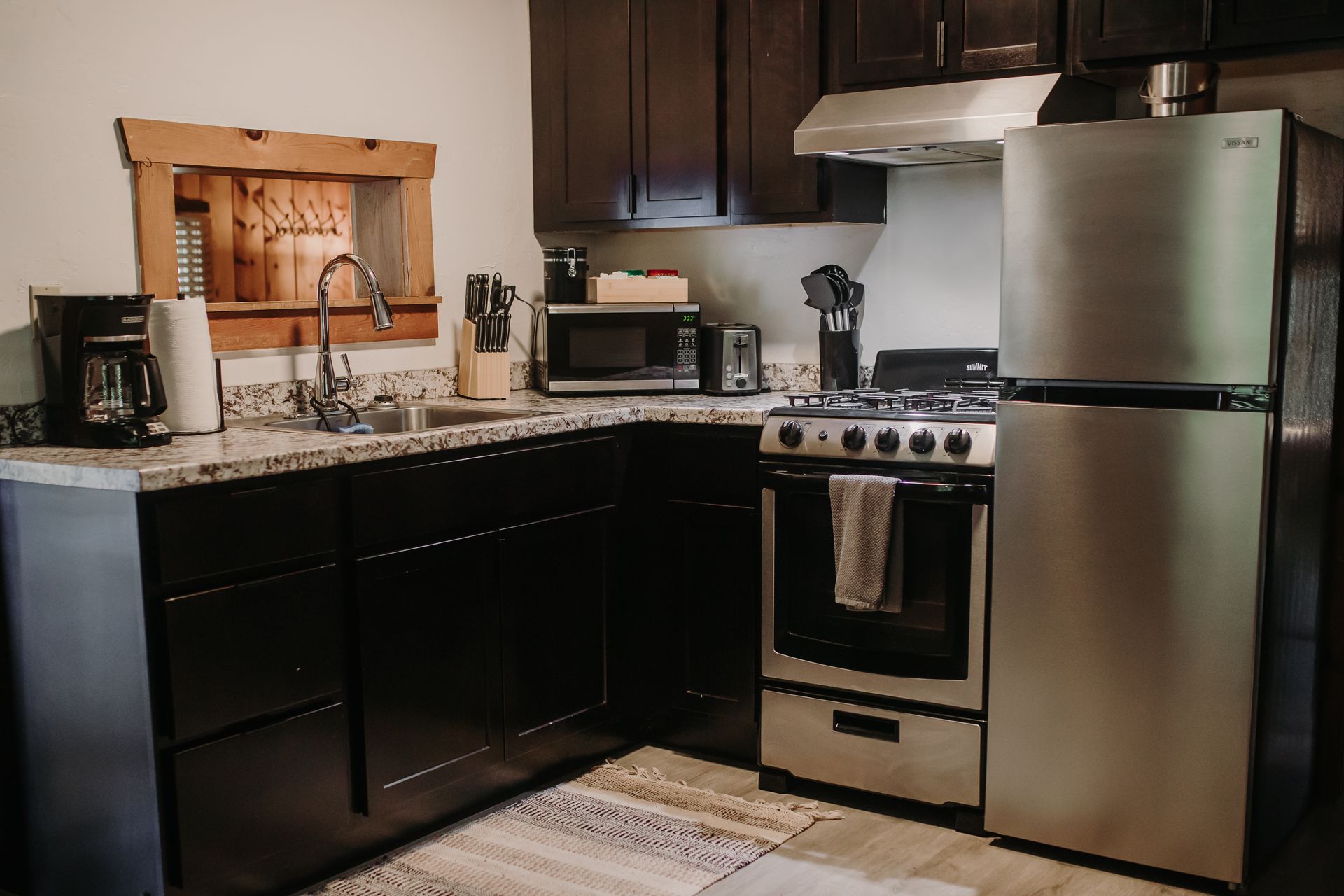A small kitchen with dark cabinets, stainless steel appliances, and a window with a rustic wood frame.