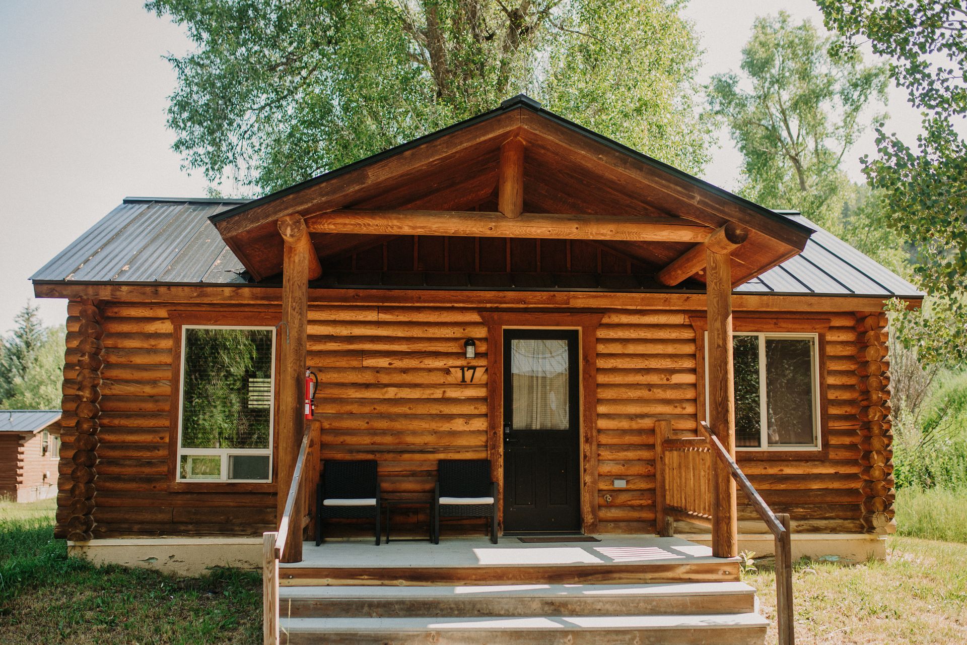 Log cabin with wooden porch, black door, two chairs, and windows.