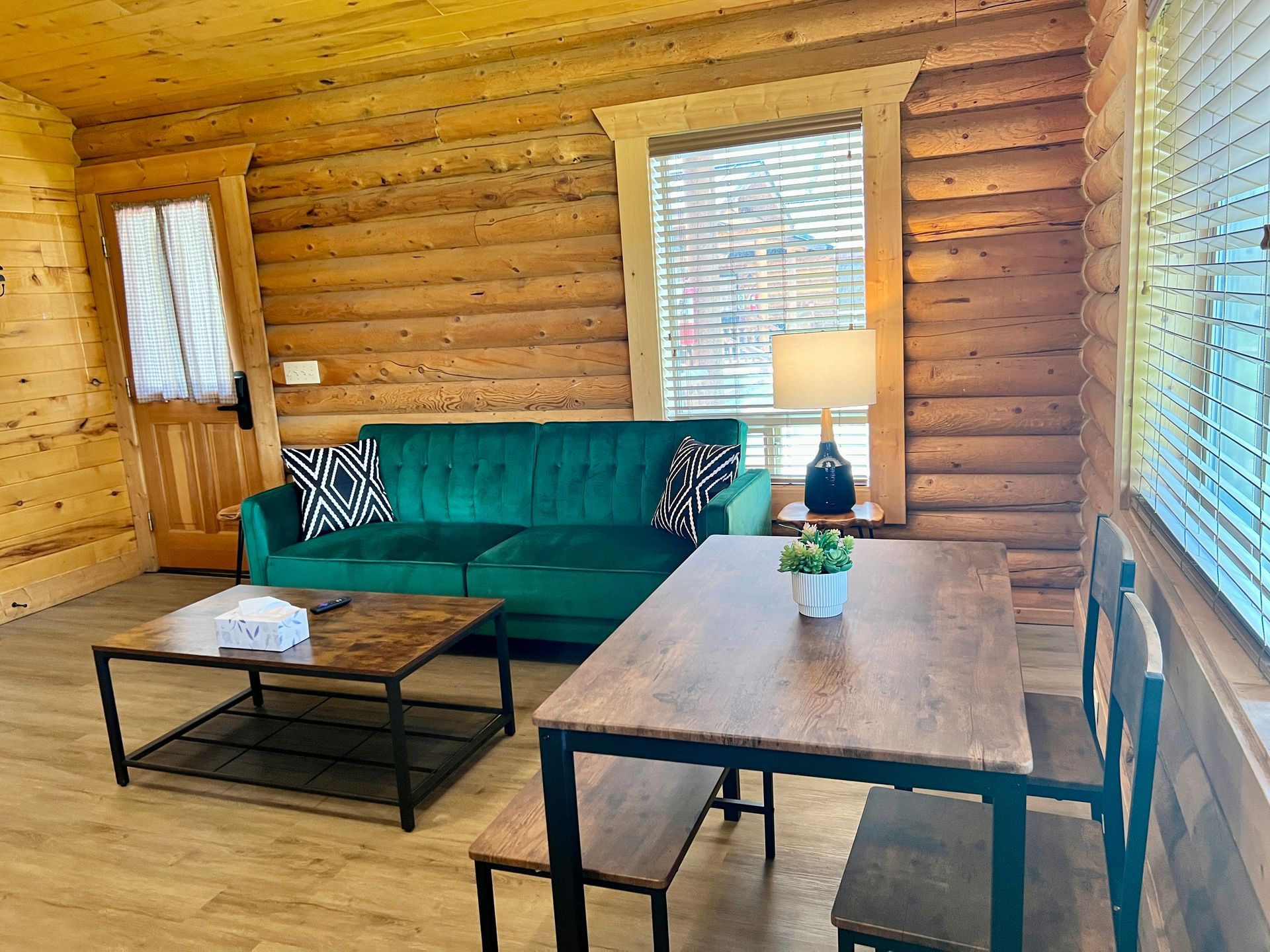 Interior of a log cabin with green couch, wooden table and chairs, and a window.