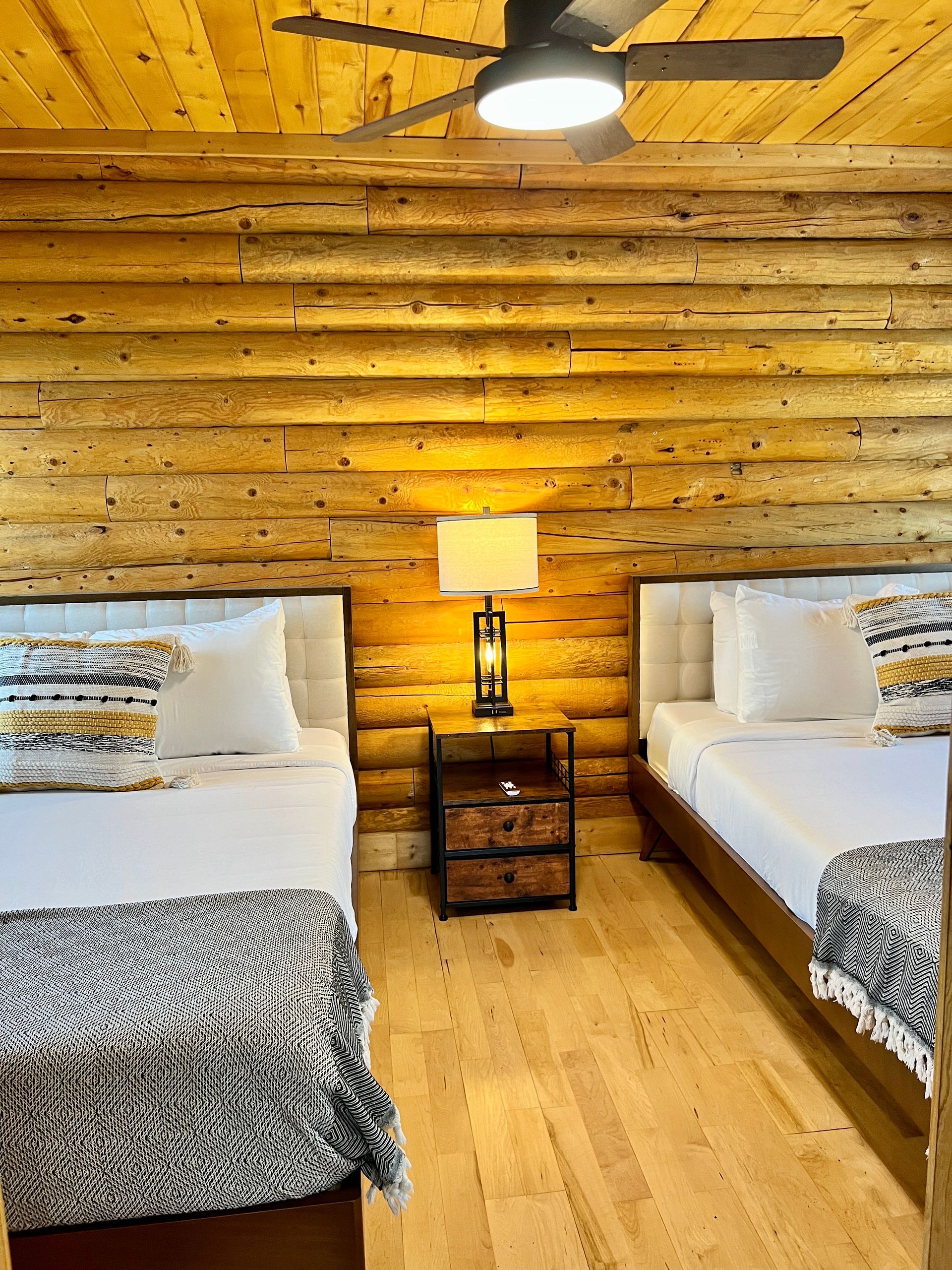 Two twin beds in a cabin room with wood walls, nightstand with lamp, and ceiling fan.