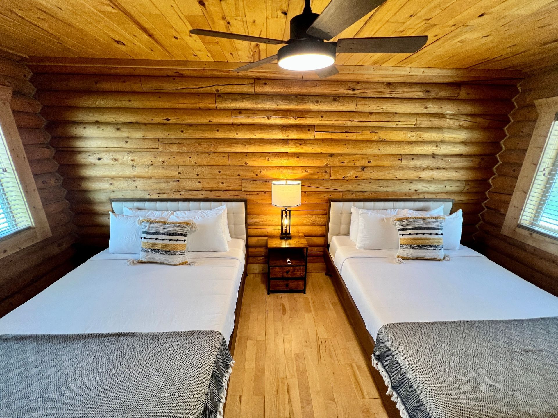 Two twin beds in a log cabin bedroom with a nightstand and ceiling fan.