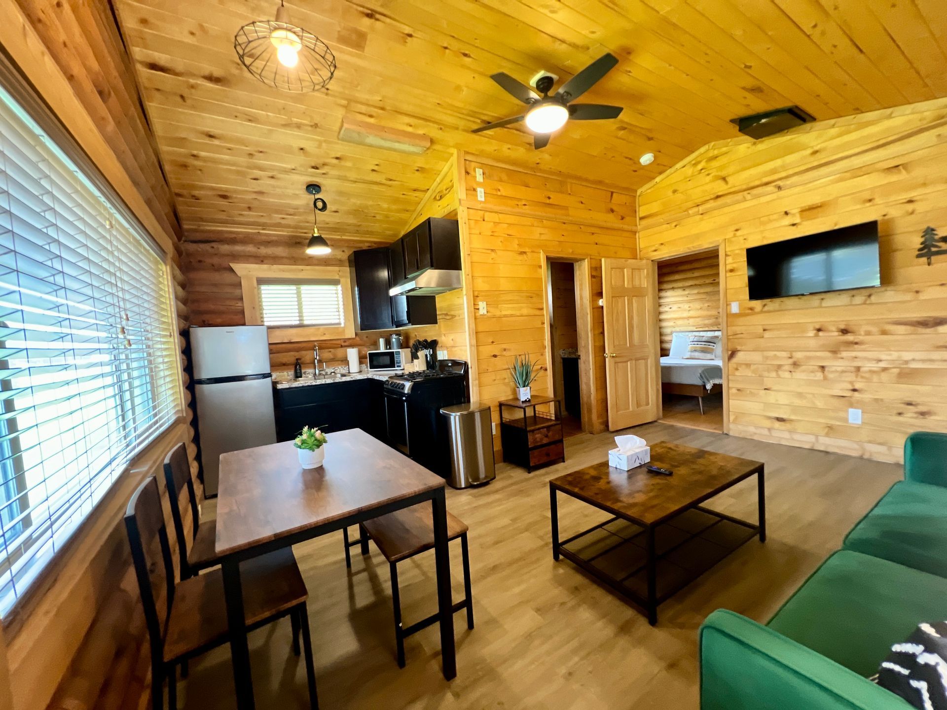 Cozy cabin interior: kitchen, living area with green couch, dining table, bedroom view. Wooden walls, ceiling.