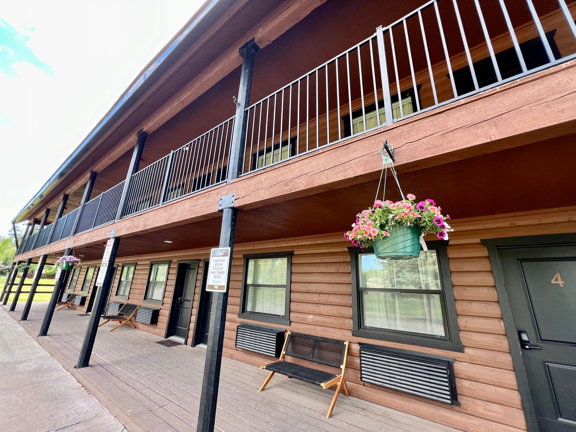 Two-story motel with brown log siding and balcony; flower basket hangs; benches and doors line the walkway.