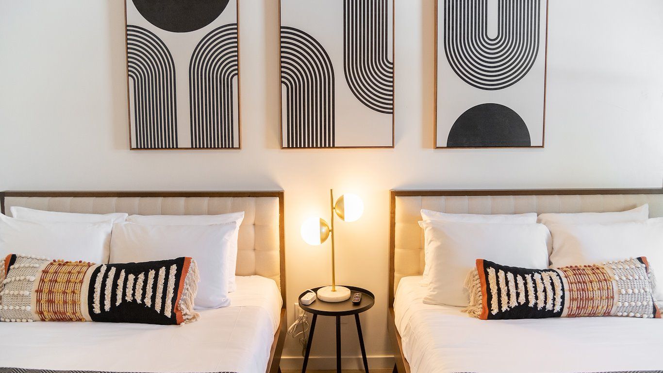 Two beds with decorative pillows under abstract black and white art, centered by a lamp on a small table.
