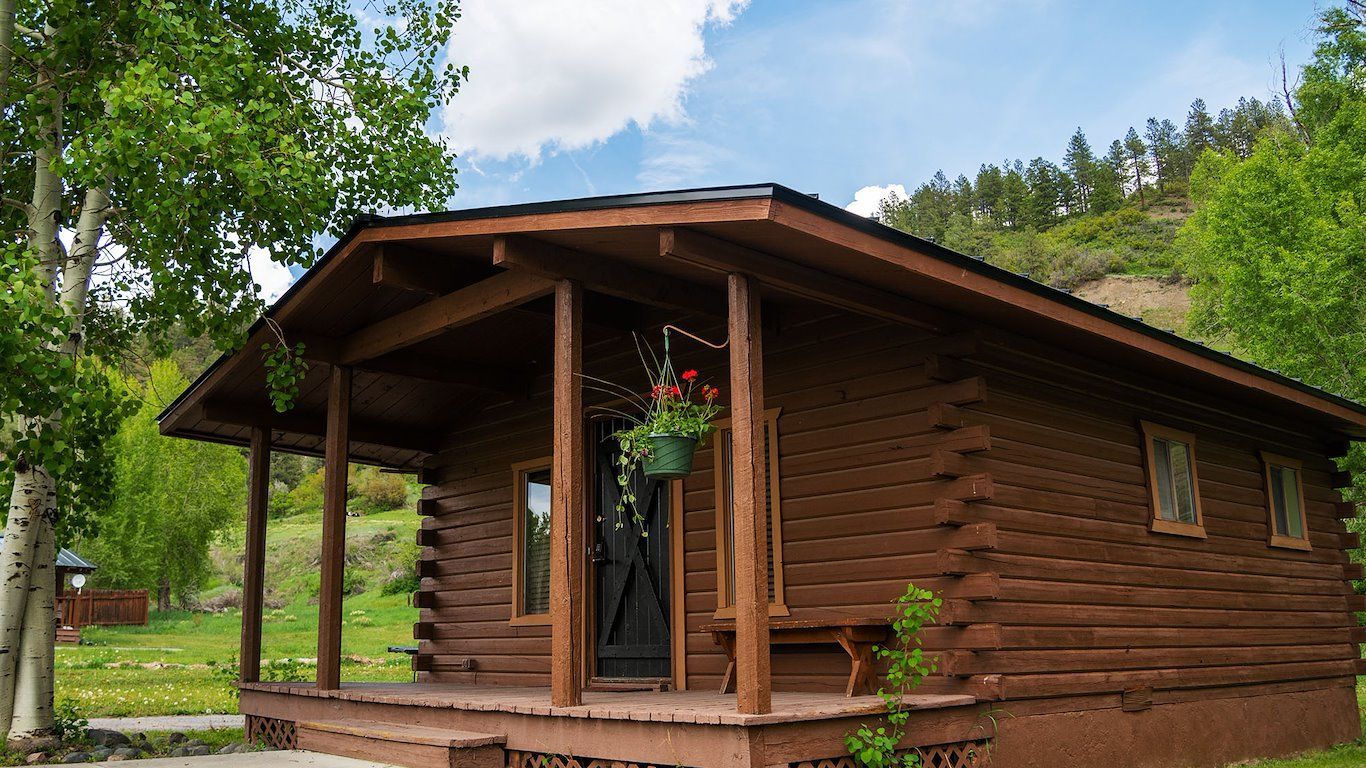 Brown log cabin with a porch, surrounded by trees and greenery.