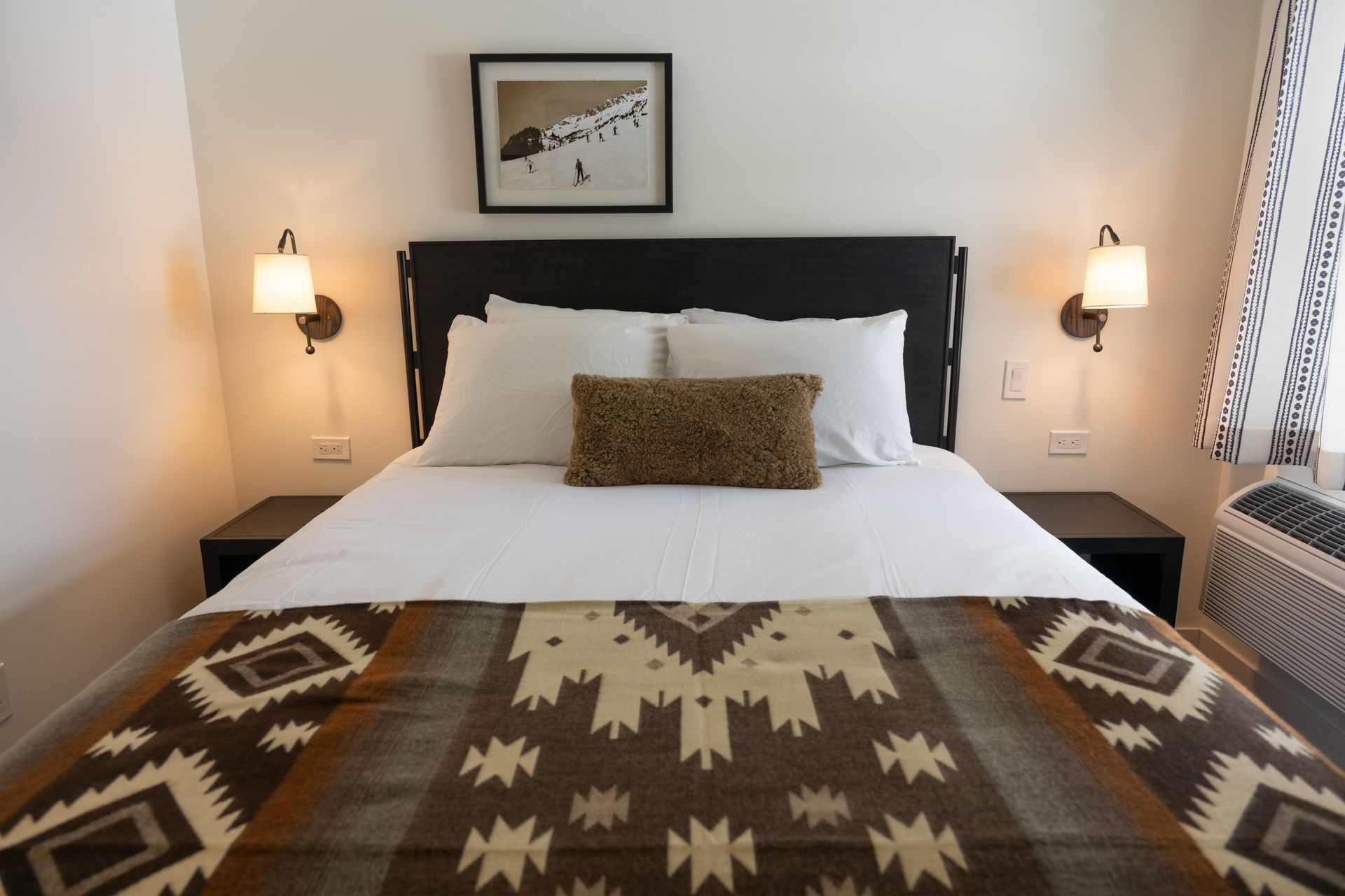 Bedroom with a double bed covered with a patterned blanket, lamps, and art above the headboard.
