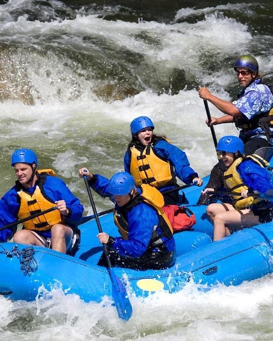 People in blue raft navigating whitewater rapids, wearing blue helmets and life vests.