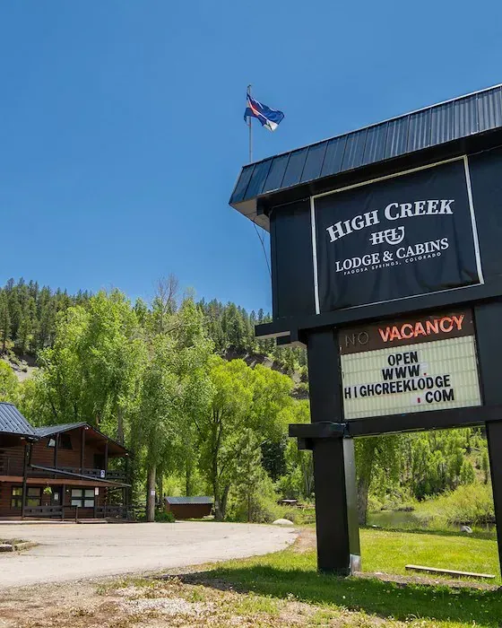 High Creek Lodge & Cabins sign with vacancy. Colorado flag, cabin, and trees under a blue sky.