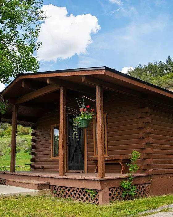 Brown log cabin with porch, hanging flowers, and a bench, set in a grassy area with a mountain in the background.
