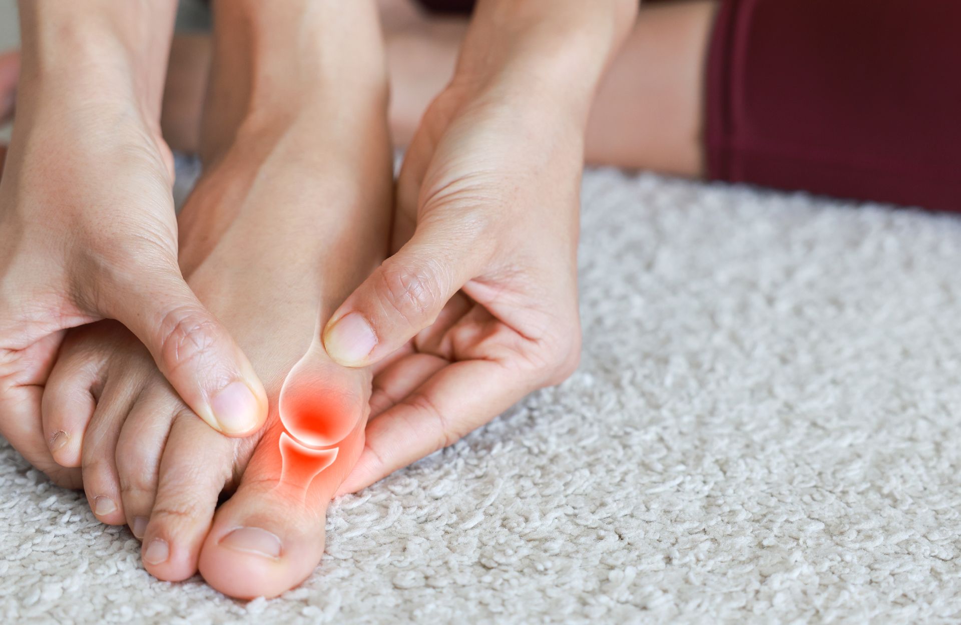 Hands massaging a painful big toe with a red highlighted joint on a carpeted floor