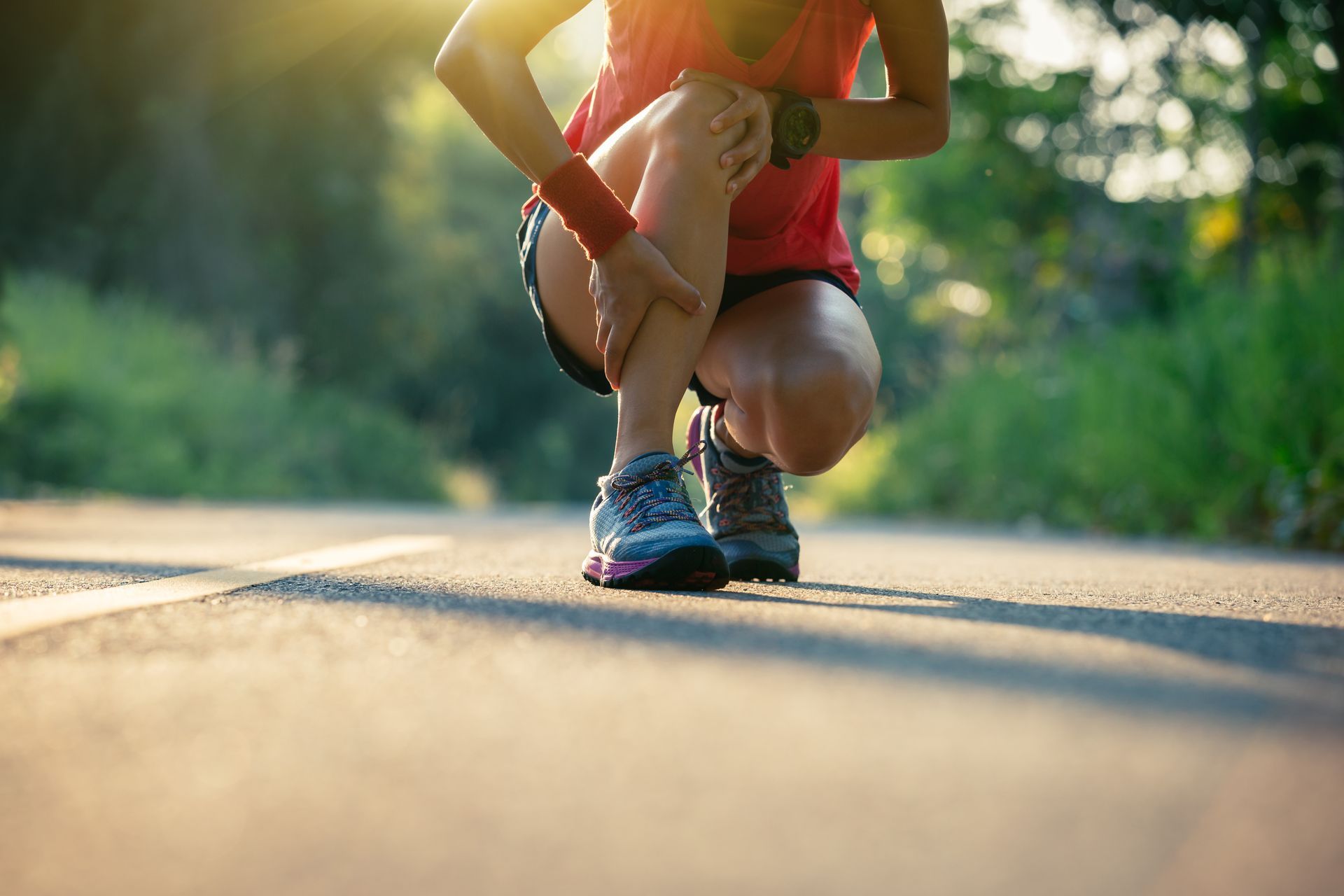 Runner tying shoelace on a sunlit road, wearing red top and blue shoes, with a blurred green background