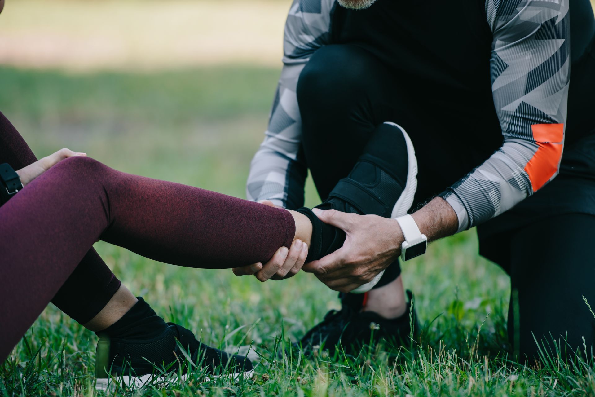 Person helping another stretch a leg on grass outdoors, with hands supporting a maroon-clad calf
