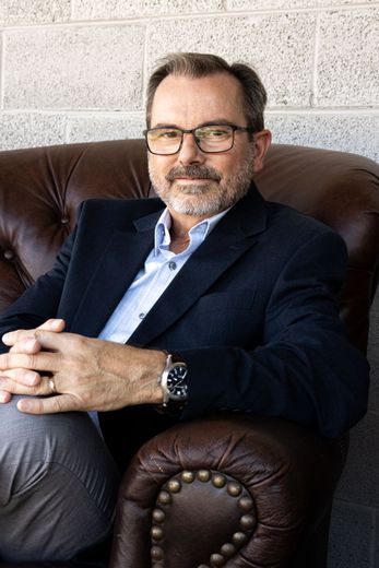 Jonathan King in a suit and glasses is sitting in a chair with his hands folded.