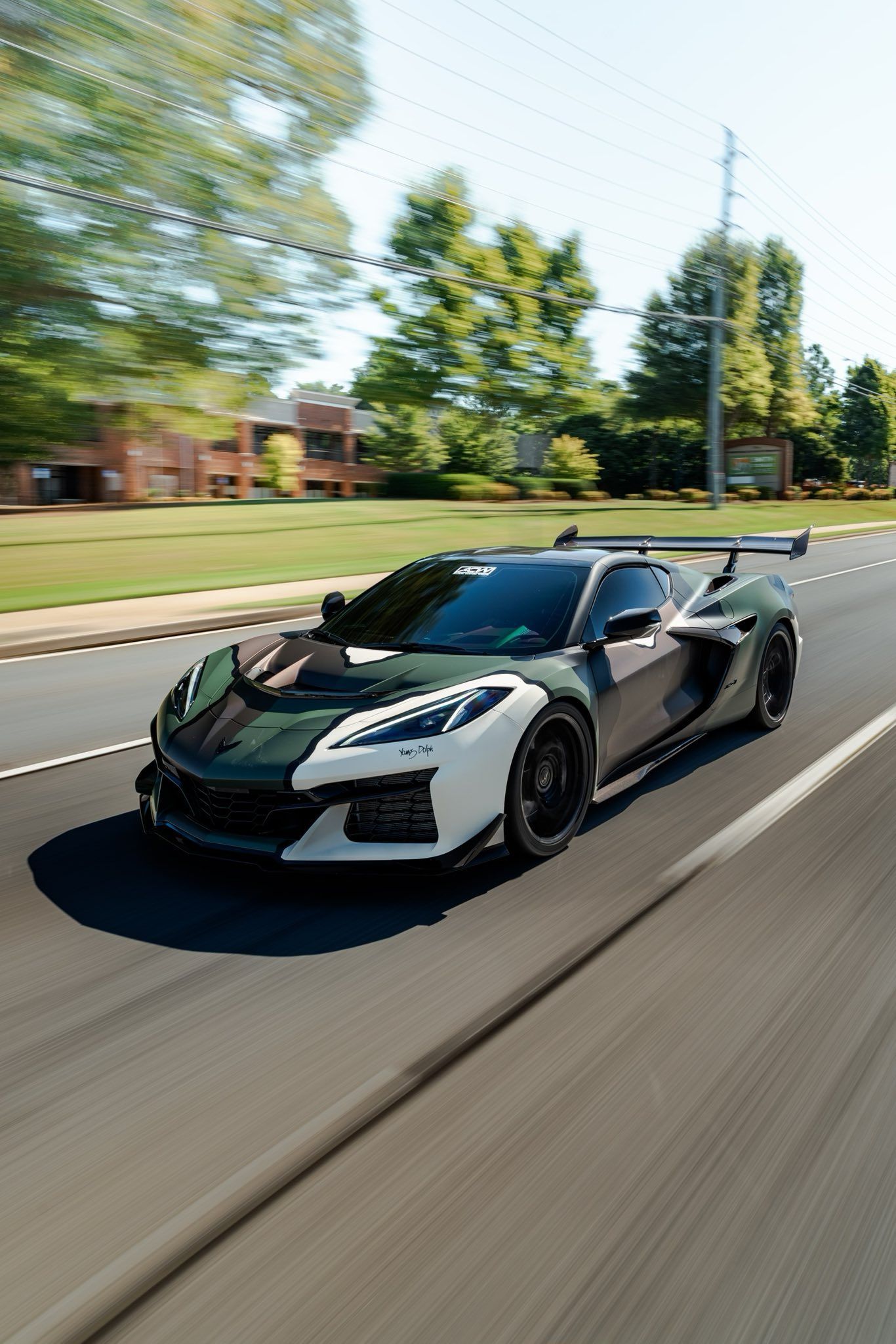 Camouflaged sports car driving on a road; white and dark green with black wheels.