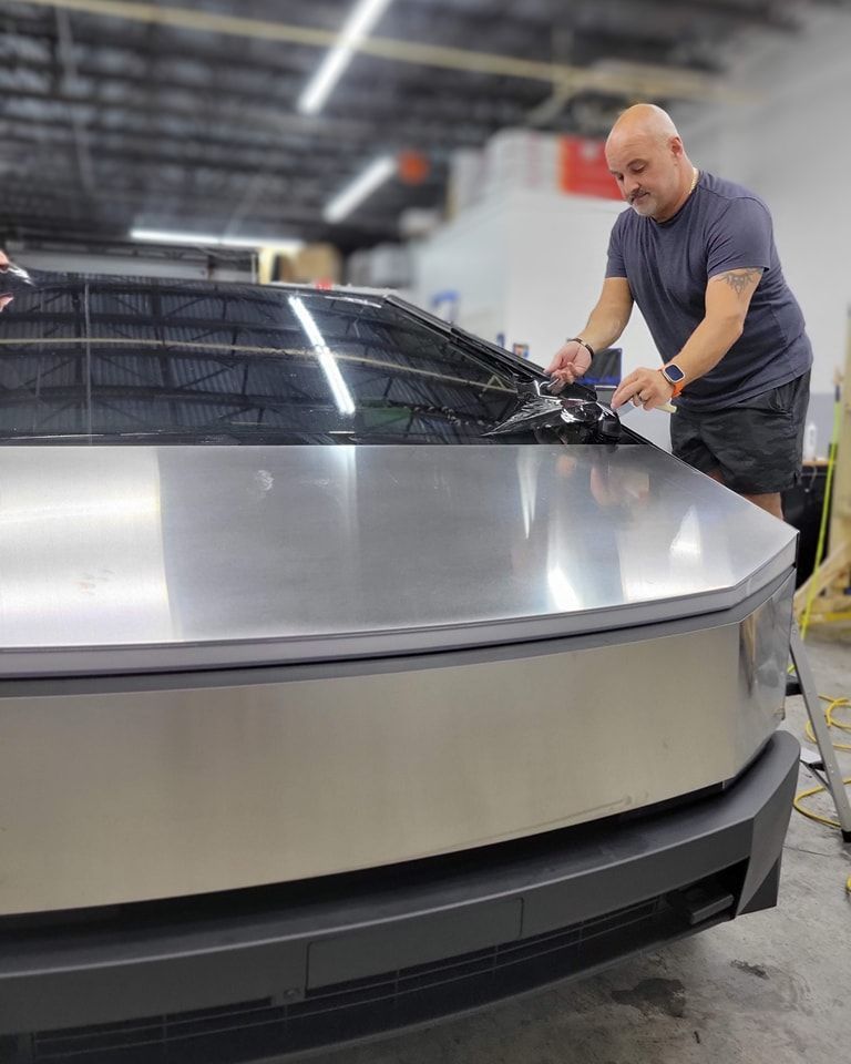 Man applying tint to the windshield of a silver Tesla Cybertruck in a workshop.