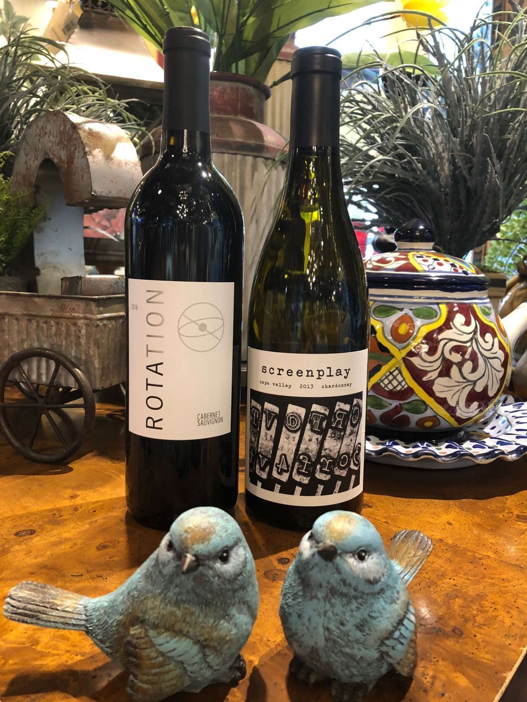Two blue bird figurines in front of two wine bottles on a wooden table with a colorful ceramic pot.