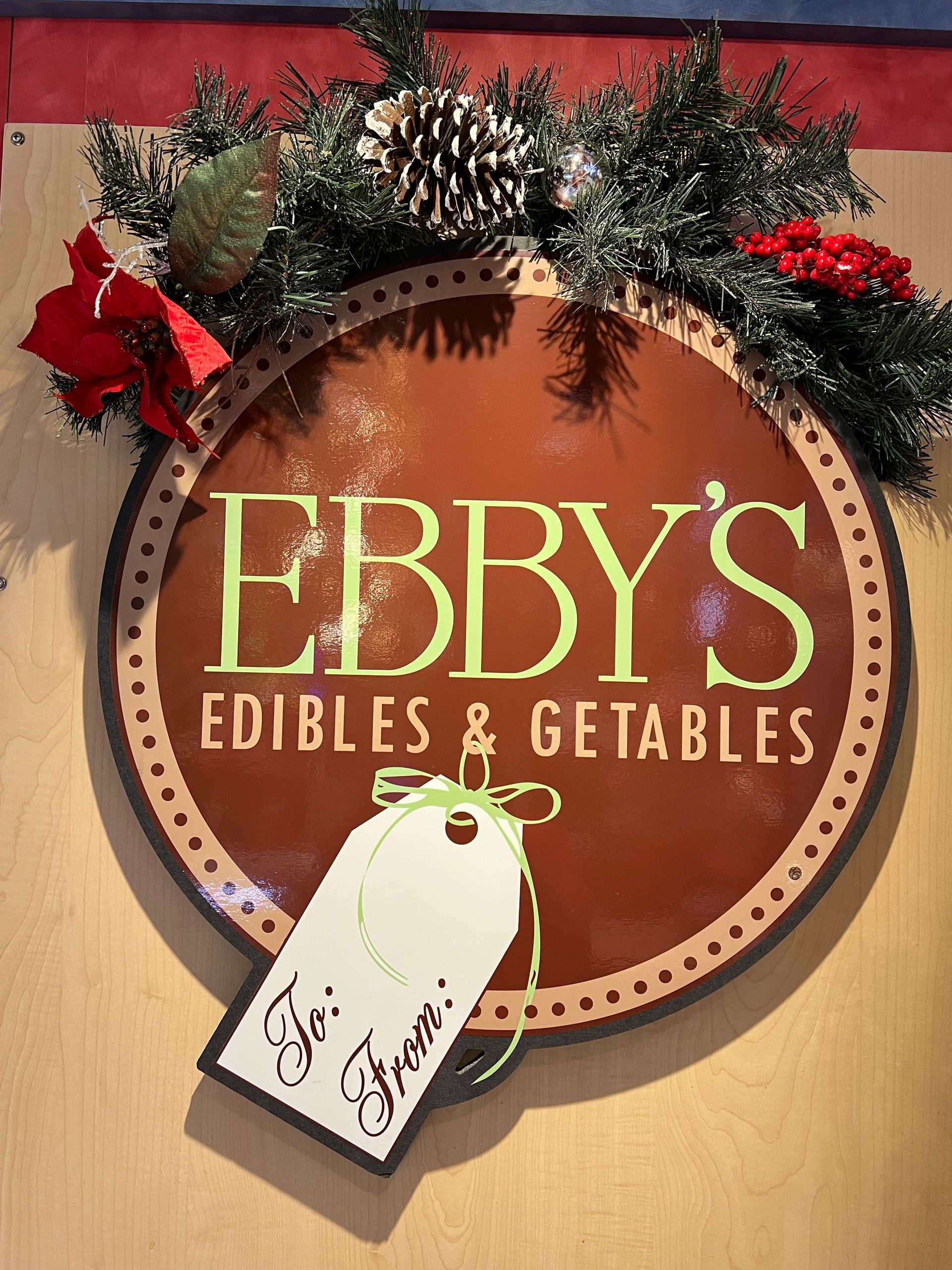 Festive “EBBY’S Edibles & Getables” wall sign with greenery, pinecones, and red bows