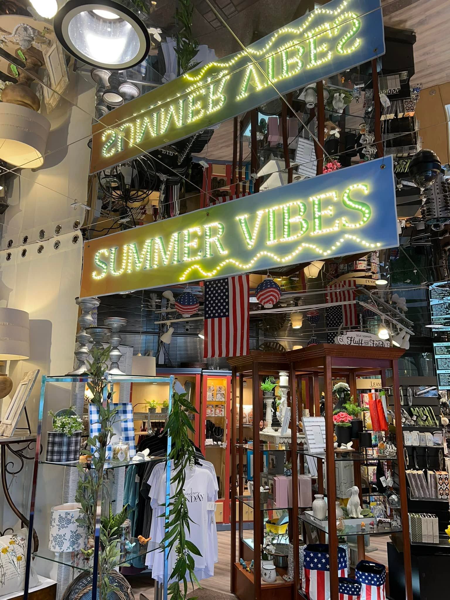 Shop entrance with neon “SUMMER VIBES” sign, hanging plants, and American flags in a colorful indoor market.