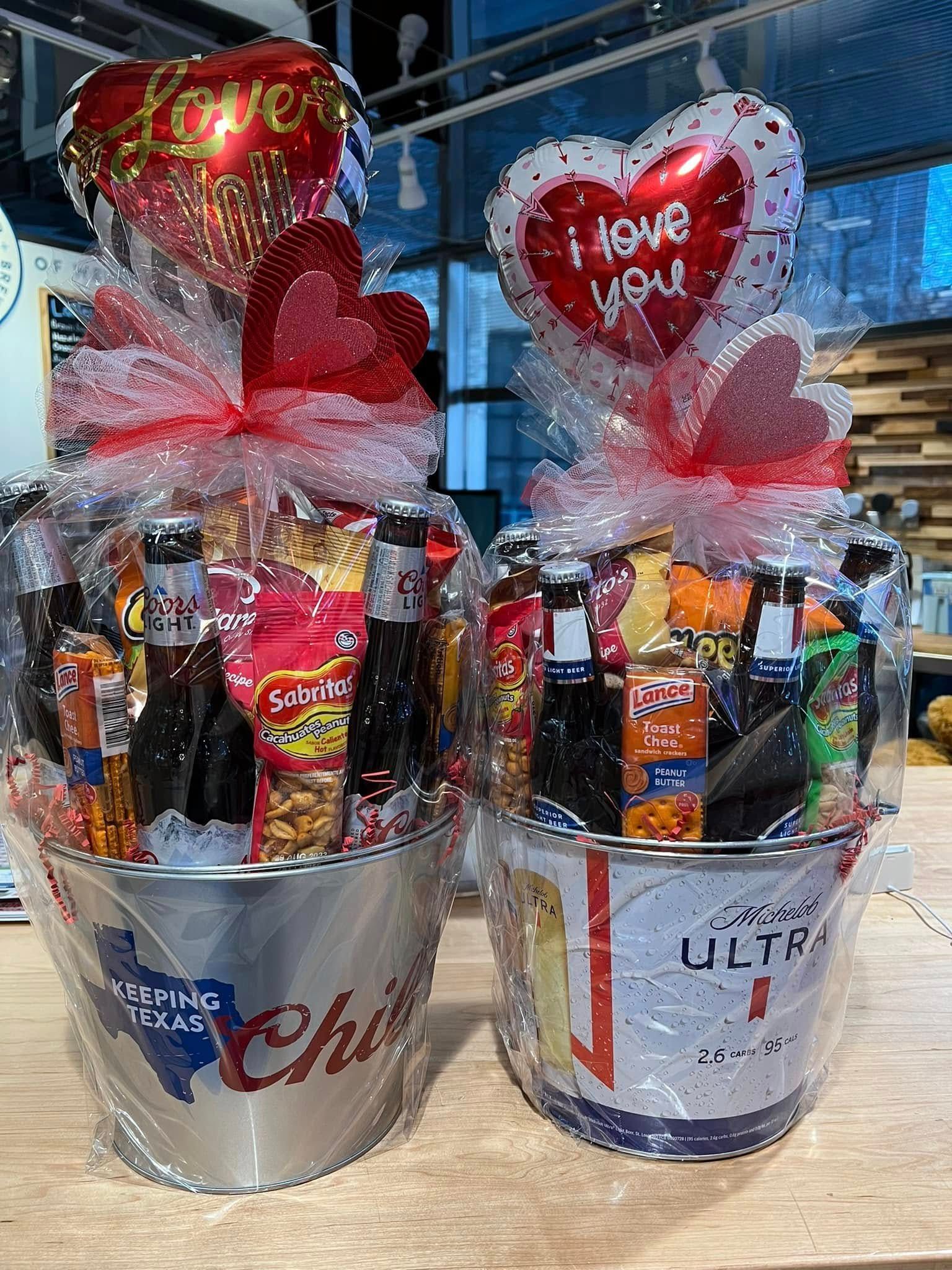 Two gift buckets filled with assorted drinks and snacks, topped with red heart balloons.