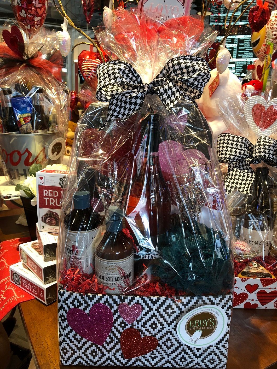 Gift basket with wine, snacks, and red heart decorations wrapped in clear cellophane.