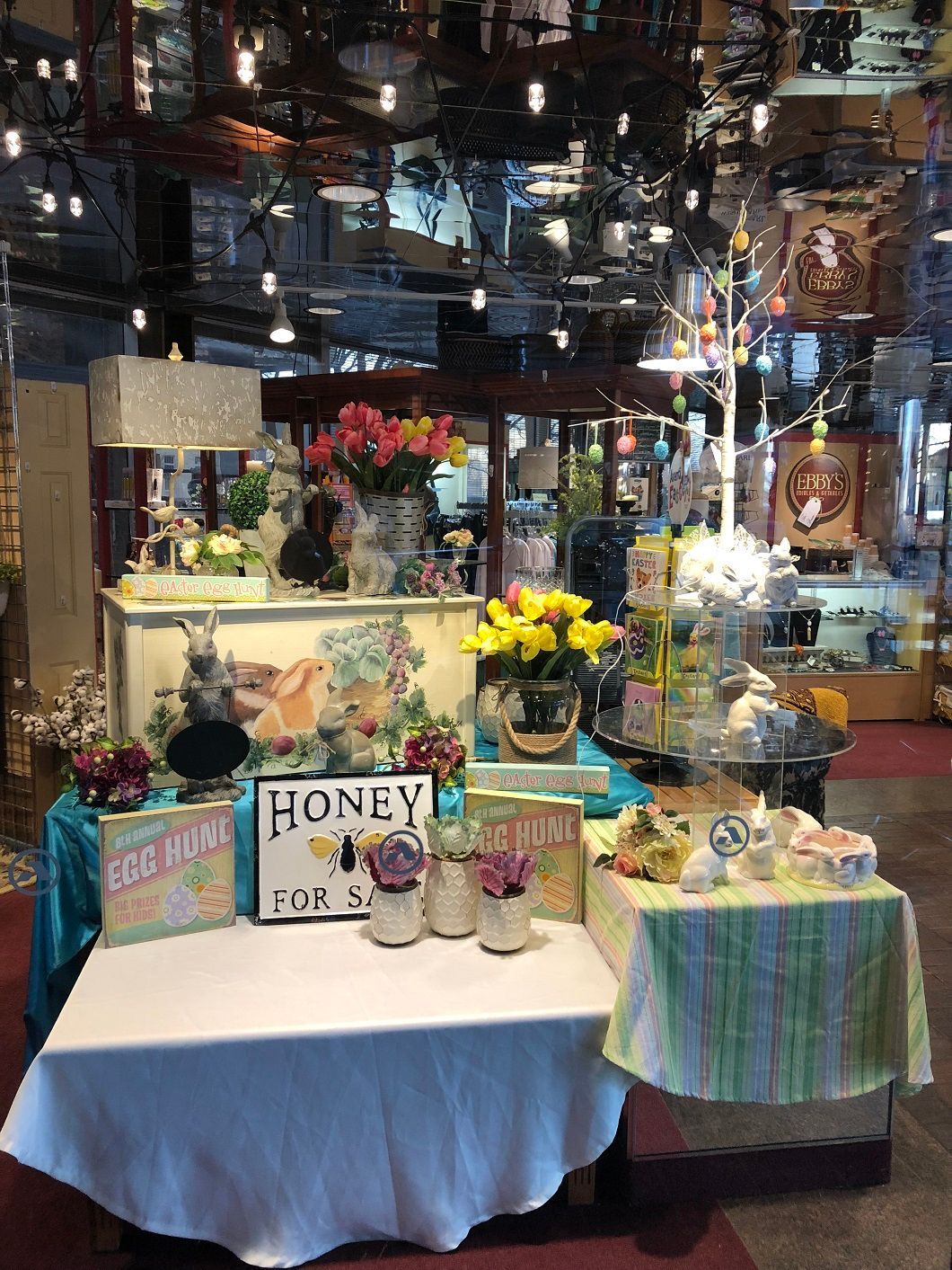 Market stall with honey jars, flowers, and colorful crafts on display under hanging lights.