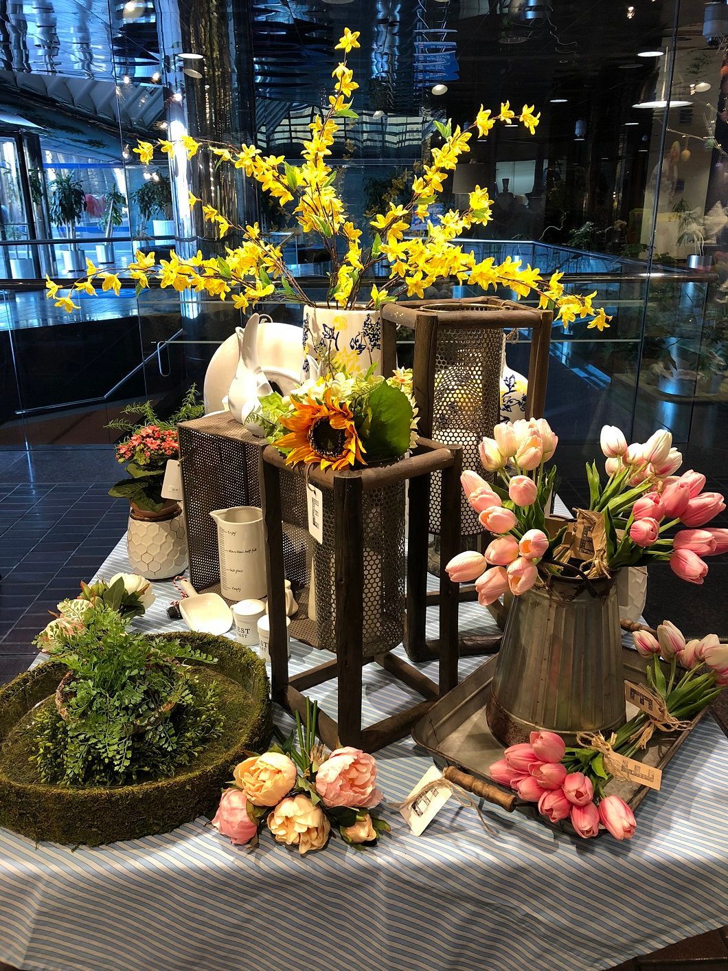 Floral display with yellow and pink blossoms, green moss, and rustic wooden stands on a gray tablecloth indoors