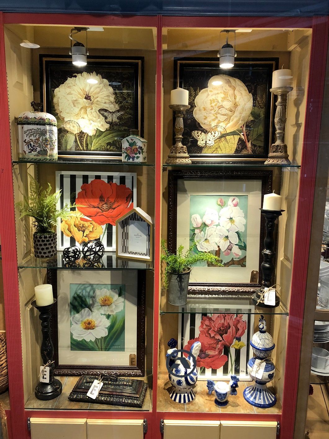 Display of framed floral artworks and decorative ceramics in a shop window, lit by warm hanging lights