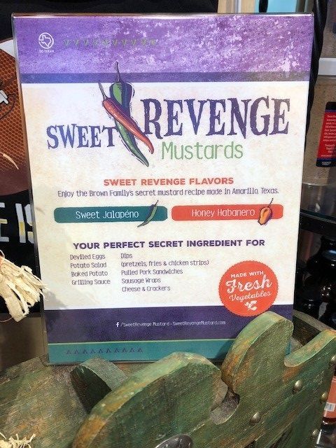Sweet Revenge mustard sign in purple and white with flavor labels and fresh basil leaves in foreground