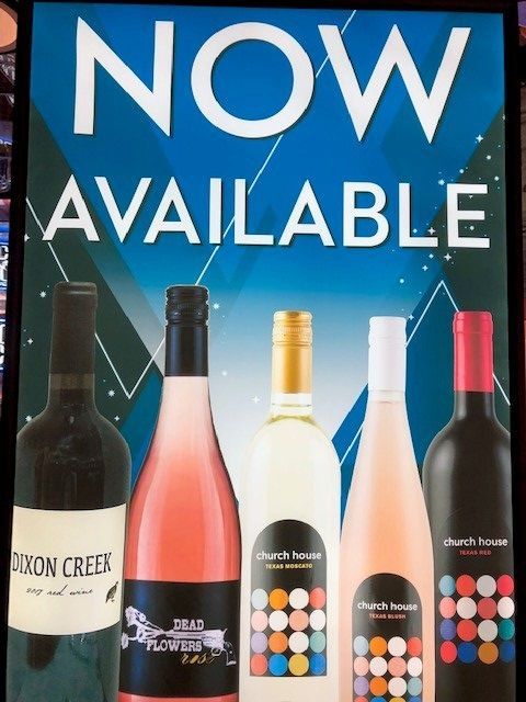 Wine bottles under a “NOW AVAILABLE” sign on a blue promotional poster