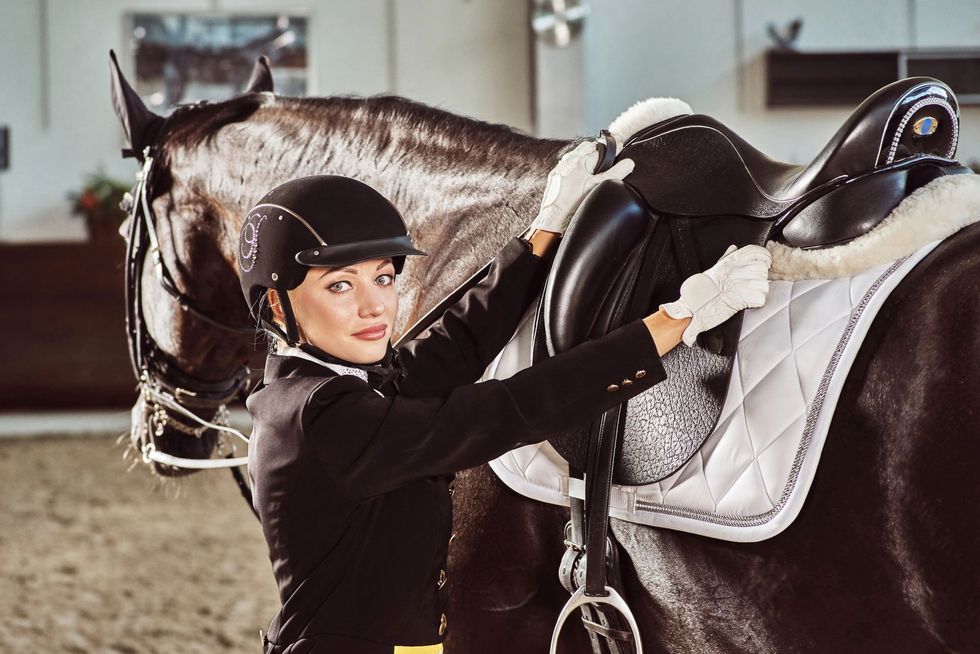 Donna in tenuta da cavallerizza che sistema la sella di un cavallo scuro in un'arena coperta.