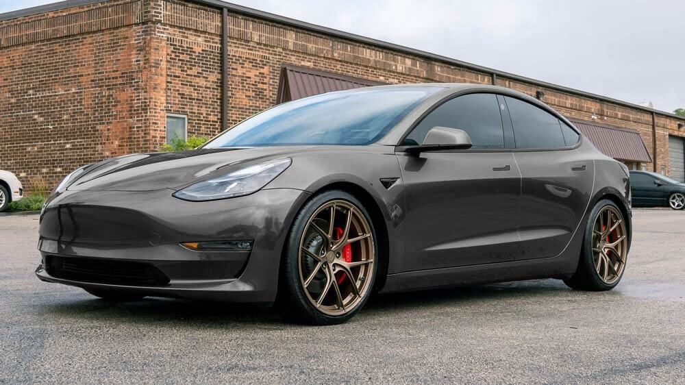 Gray Tesla Model 3 on a wet street with bronze wheels, parked in front of a brick building.