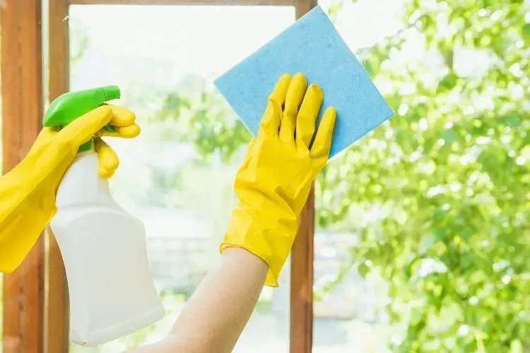 A person wearing yellow gloves is cleaning a window with a sponge and spray bottle.