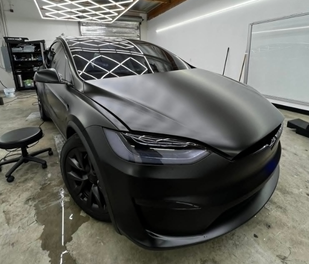 Matte black Tesla Model X in a garage, with a stool and office chair nearby