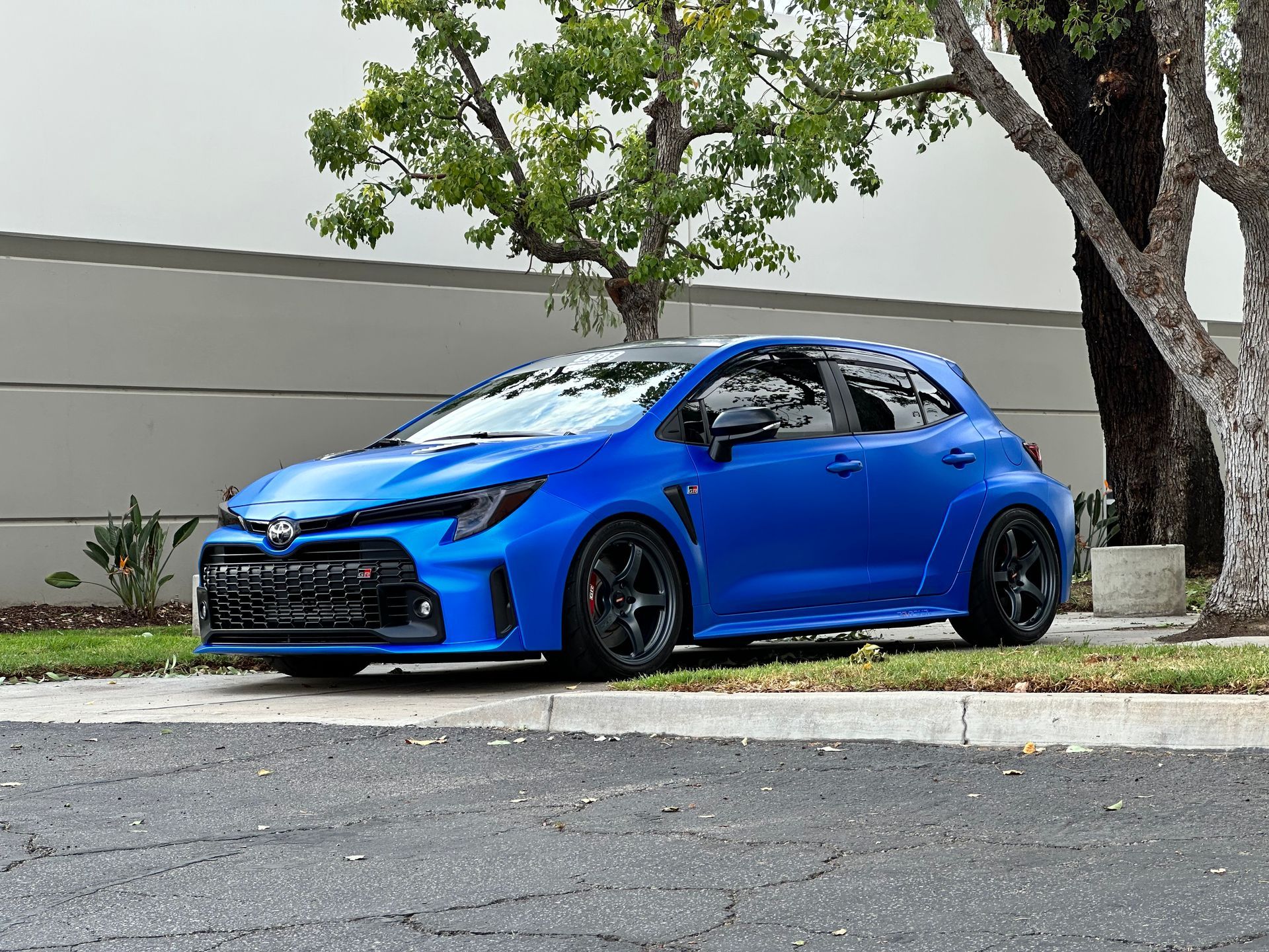 Blue hatchback car parked by a curb beside trees and a gray wall