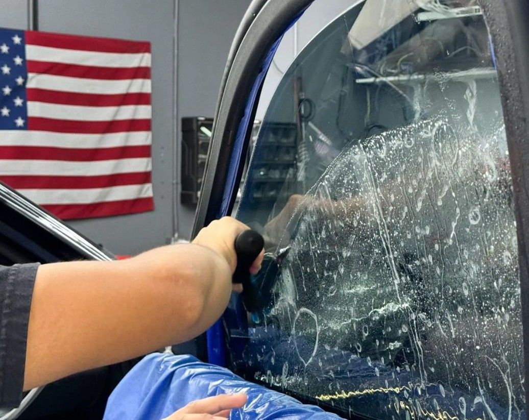 Hand breaking a car window with a tool; shattered glass and U.S. flag in background.