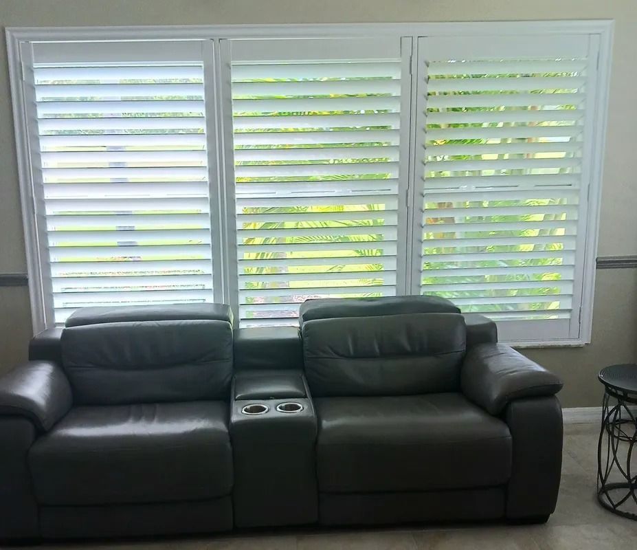 Black leather sofa in front of white window shutters with daylight filtering through.