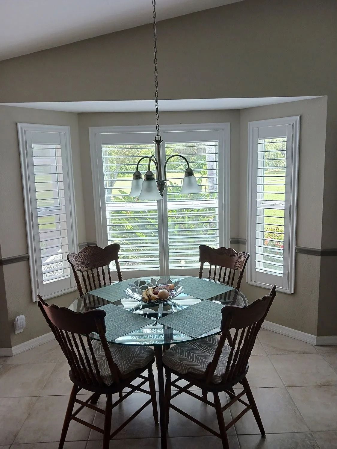 Dining nook with round glass table, four chairs, pendant light, and bay windows with blinds.
