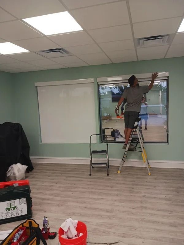 Person on a ladder painting around a window in a room with mint walls and a whiteboard