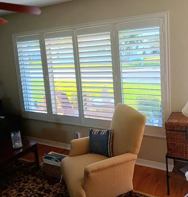 Living room with beige armchair by a large window with white shutters, wood floor, and a side table.