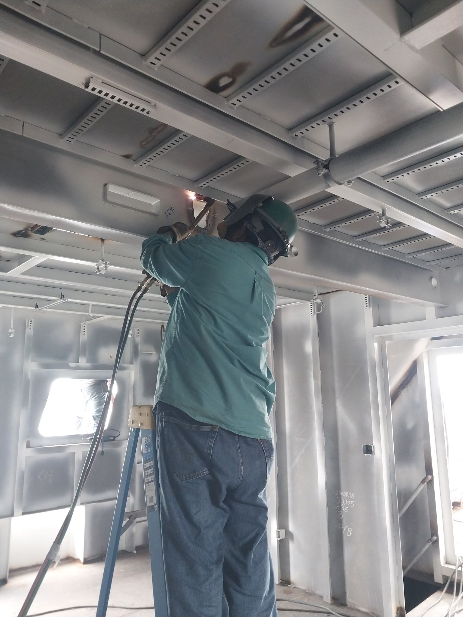 Worker in green shirt welding overhead on a metal ceiling inside a steel structure.